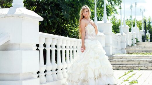 Beautiful bride in an elegant white ball gown posing outdoors on a sunny day beside a white balustrade and garden backdrop.