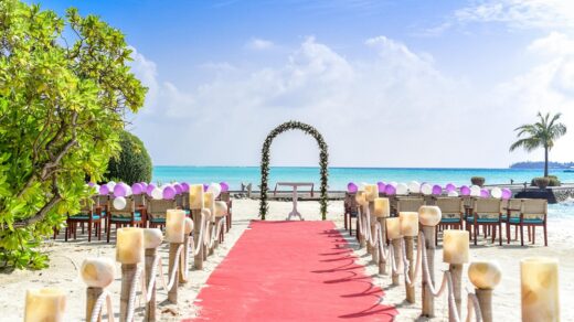 Beach wedding venue with red carpet aisle, floral arch, and chairs decorated with purple and white balloons under a clear blue sky
