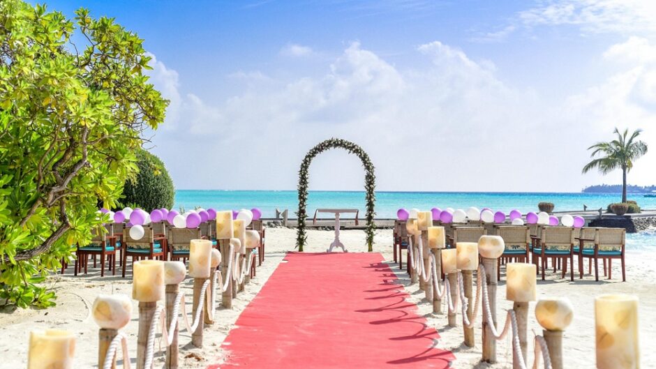 Beach wedding venue with red carpet aisle, floral arch, and chairs decorated with purple and white balloons under a clear blue sky