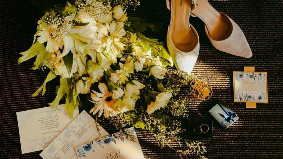 Elegant flat lay of wedding invitations, bridal shoes, bouquet of white flowers, gold rings, and gift boxes arranged beautifully on a textured surface.
